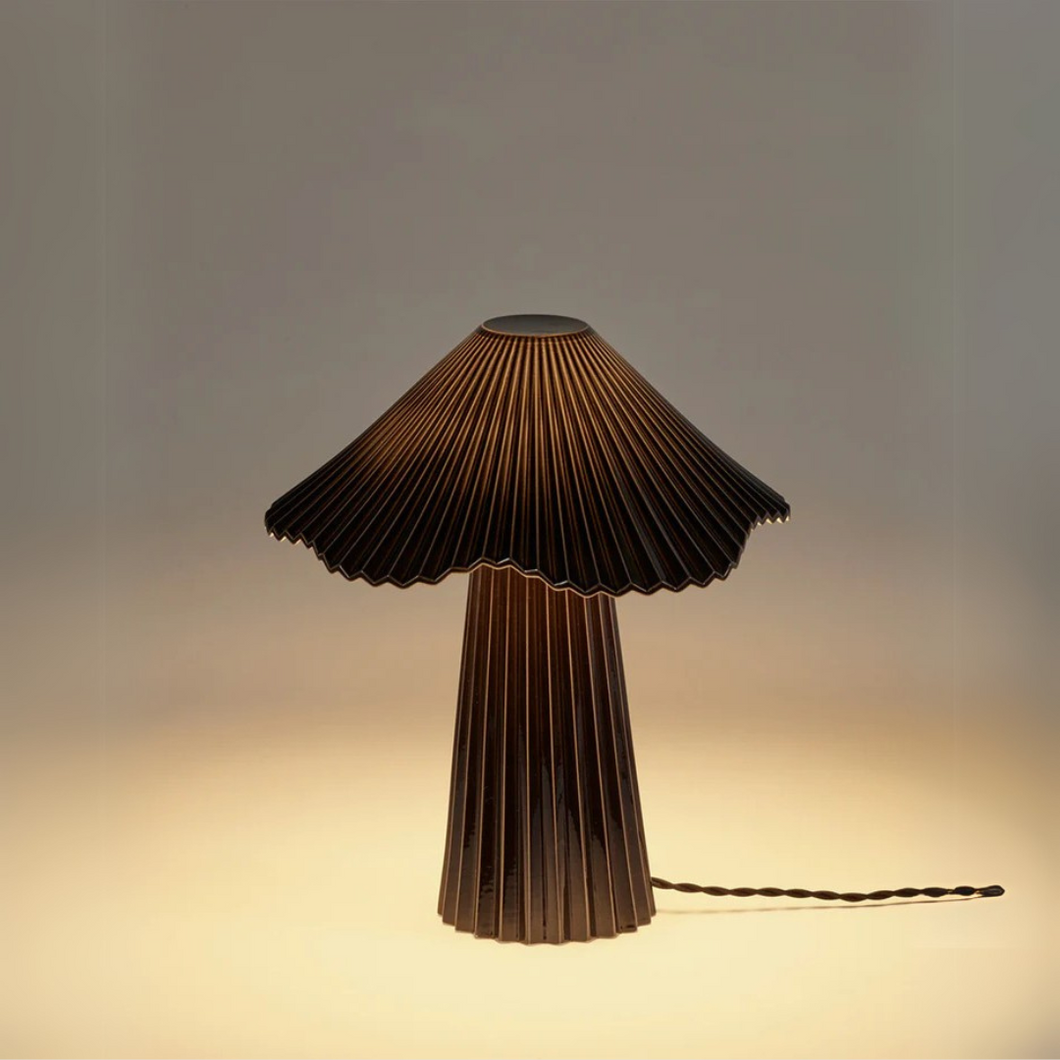 Schwarze Tischlampe „Uala“ aus feinem Porzellan mit plissiertem Schirm, Design von Sebastian Herkner für Serax. Beleuchtet, auf hellem Hintergrund. Black porcelain table lamp “Uala” with pleated shade